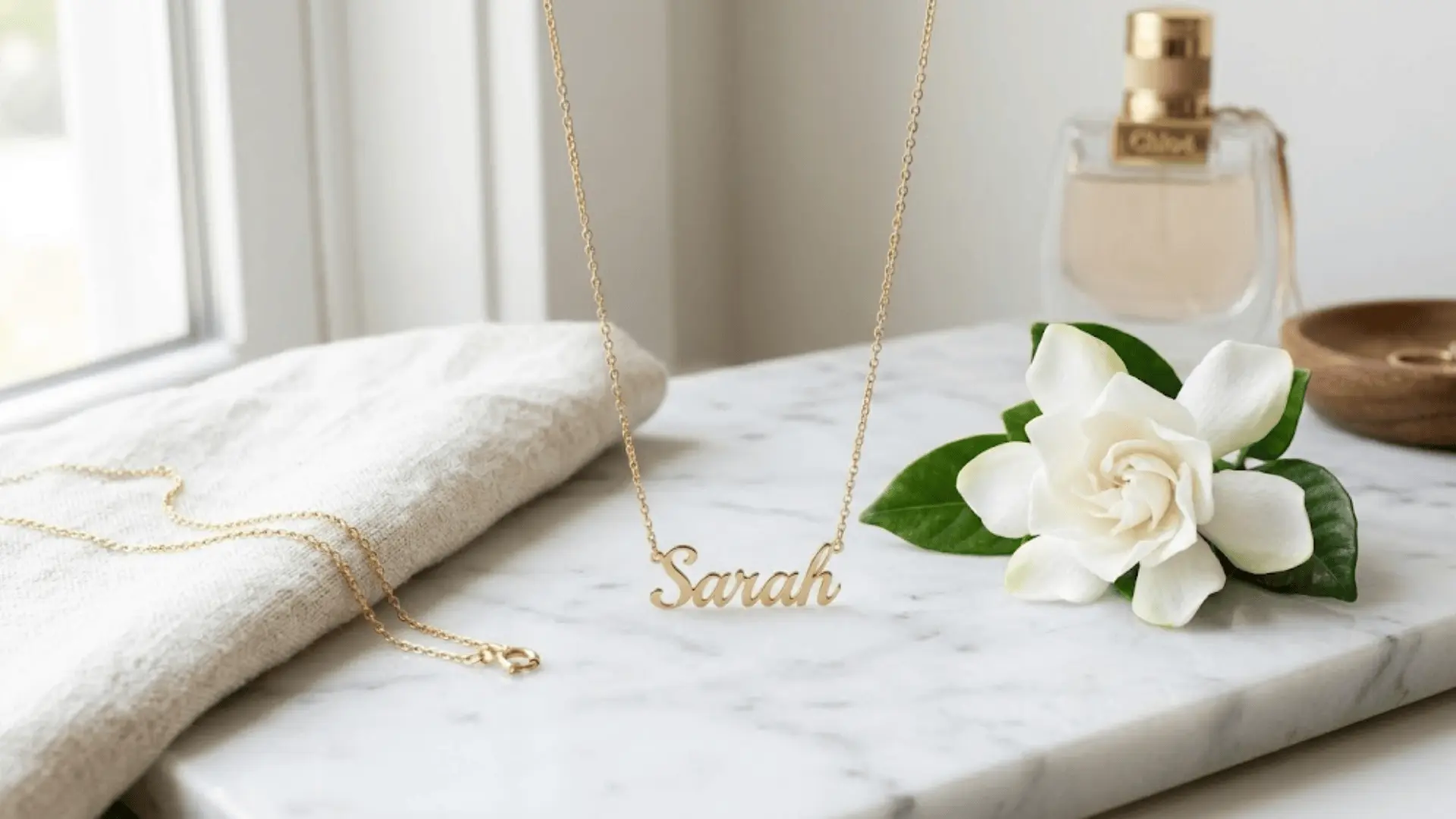 delicate gold name necklace on white marble with a gardenia and cream linen in soft natural light
