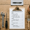 Flat lay of home maintenance checklist on clipboard with gloves, wrench, screwdriver, brush, tape, and plant on wooden table