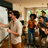 Group playing Pictionary indoors, one person drawing on whiteboard while teammates cheer, laugh, and guess in a lively setting