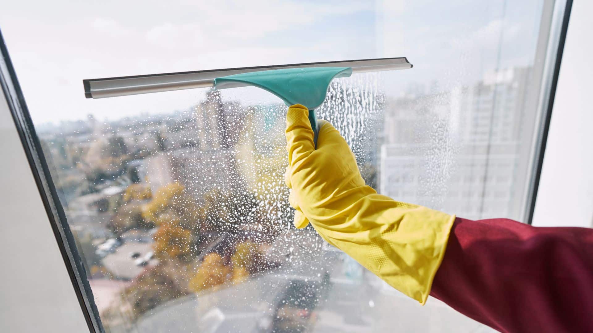 Hand using a squeegee to clean a window with a DIY glass cleaner, removing soap and water for a clear, streak-free finish