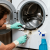 Man wearing gloves scrubbing mold from washing machine gasket, with cleaning supplies nearby in a modern laundry room.