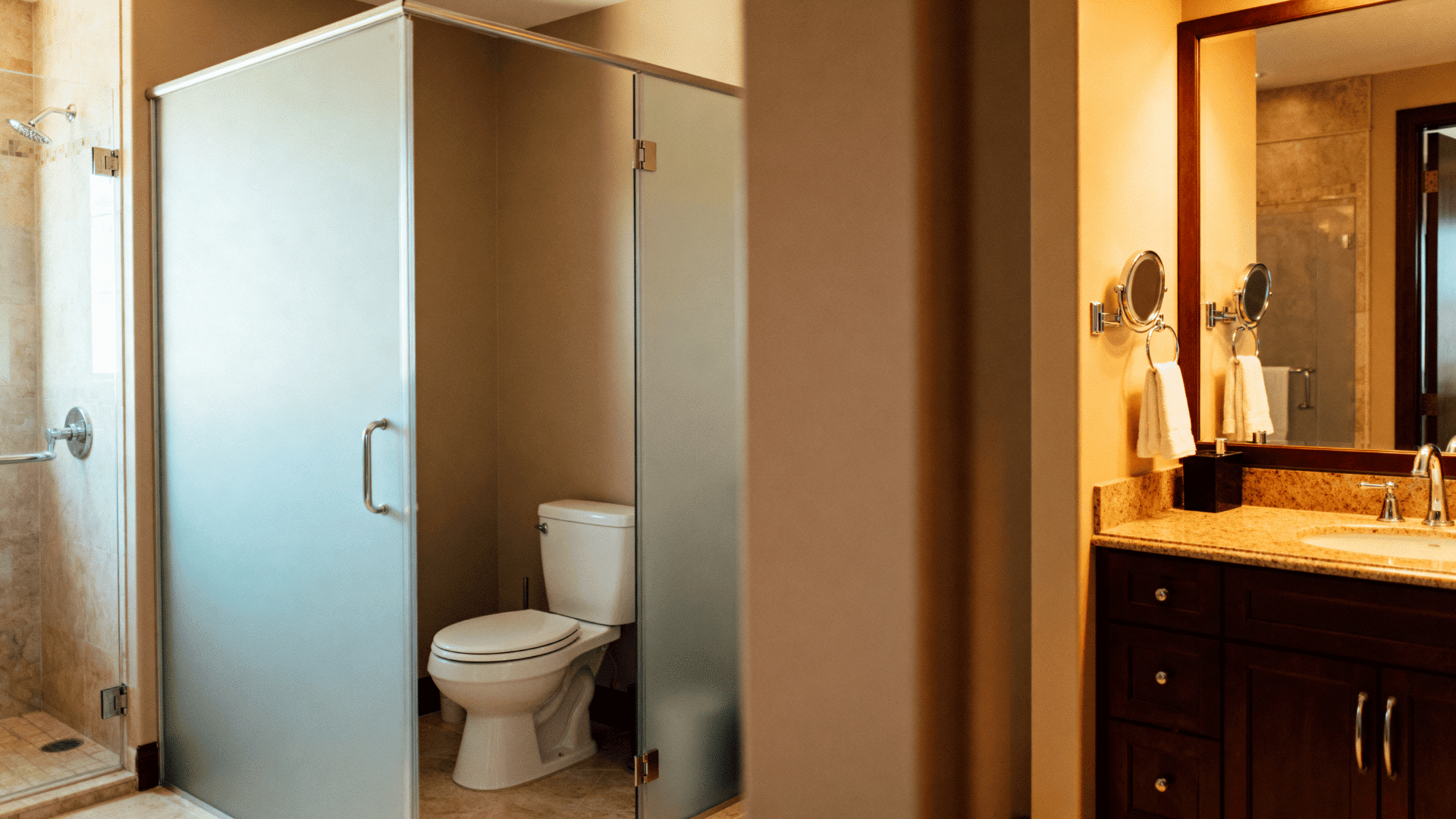 Modern bathroom with glass-enclosed toilet, walk-in shower, granite vanity, warm lighting, wood cabinetry, and functional layout design