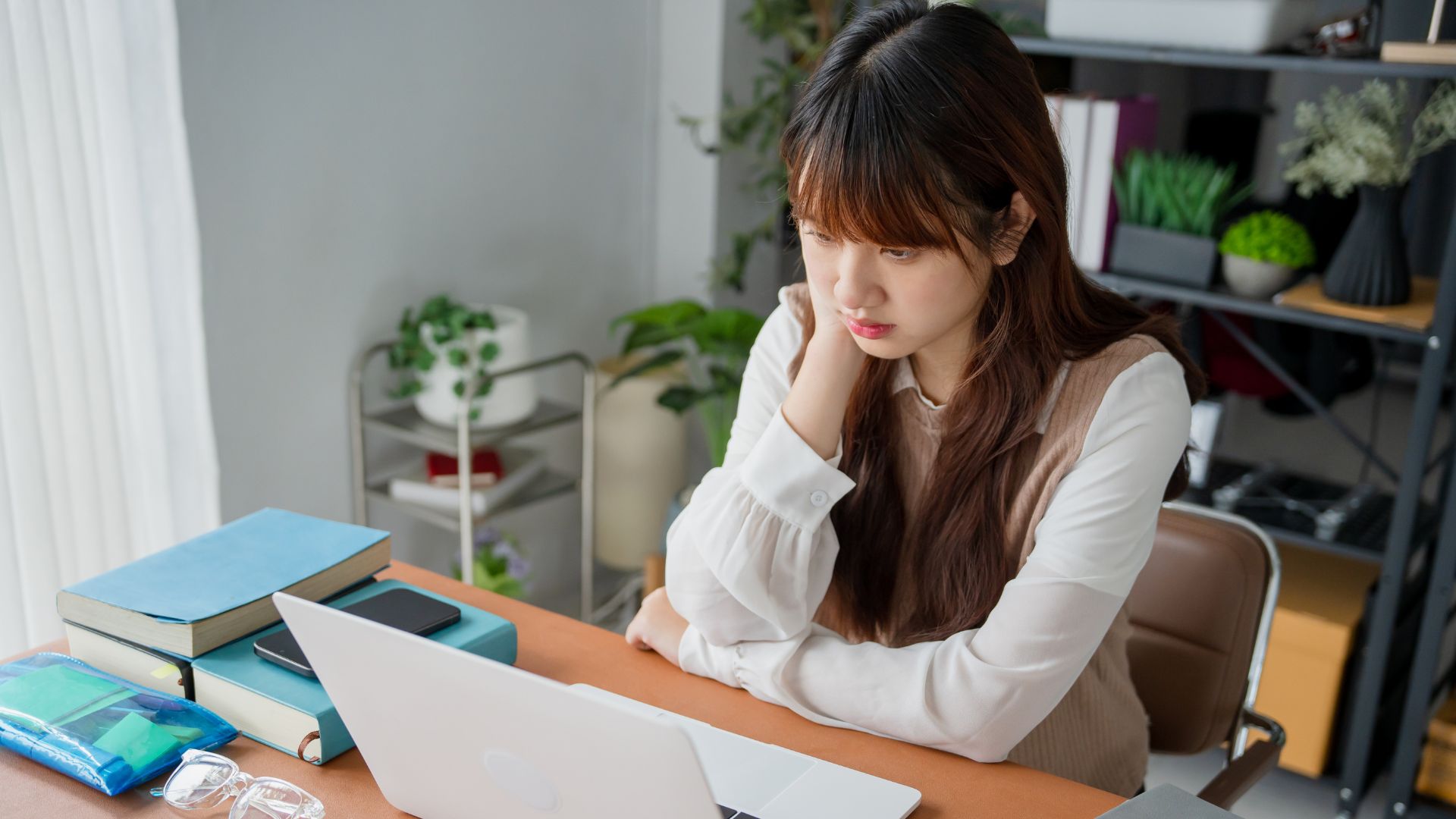 Student looking stressed while attending a virtual classrooms session on laptop at home desk with books, highlighting online learning challenges