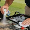 Person cleaning car battery with spray and cloth outdoors on a sunny day