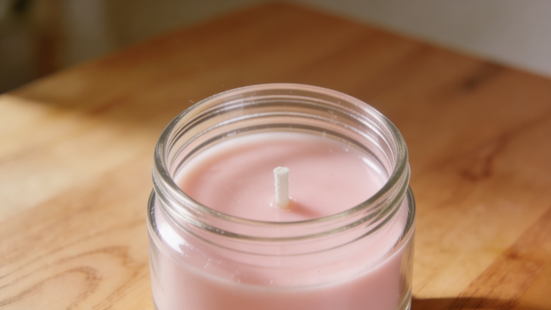A clear glass jar filled with pale pink wax cooling and hardening at room temperature on a wooden table, with a centered wick standing upright.