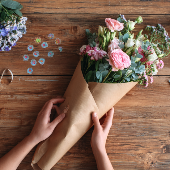 Handmade paper flower bouquet wrapped in kraft paper and ribbon surrounded by colorful origami paper and craft supplies on a white wooden table
