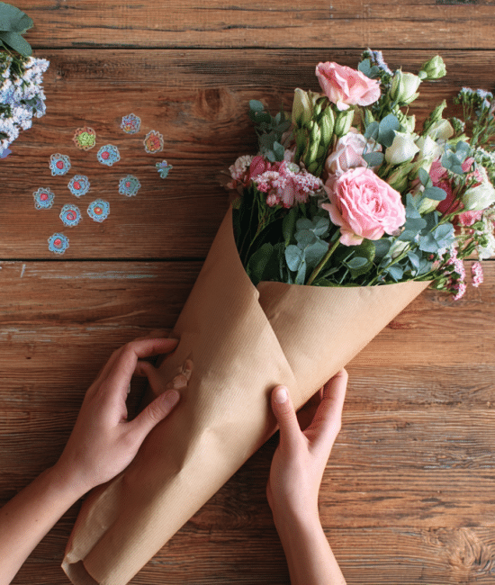 Handmade paper flower bouquet wrapped in kraft paper and ribbon surrounded by colorful origami paper and craft supplies on a white wooden table