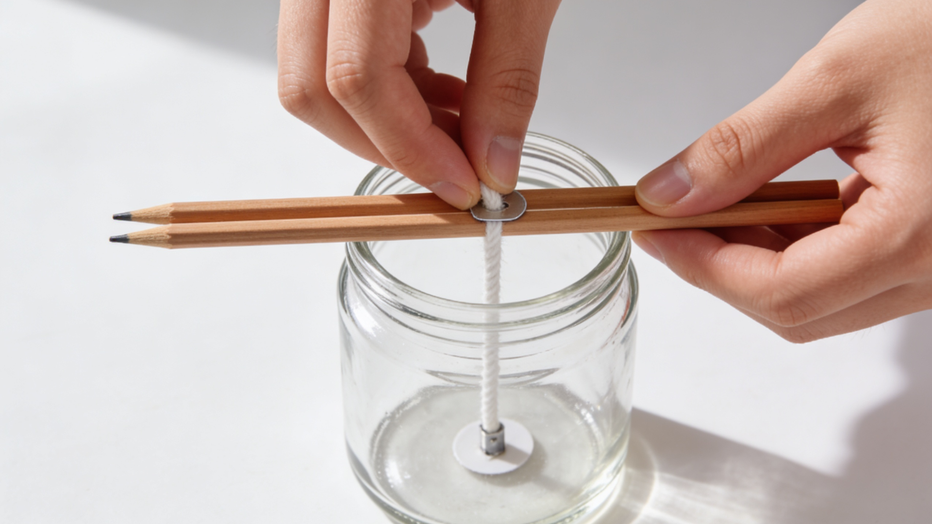 Hands placing a cotton wick at the bottom of a clear glass jar, with two pencils laid across the top to keep the wick centered and upright.