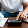 Person calculating mortgage payments with model house, calculator, and laptop on desk home.