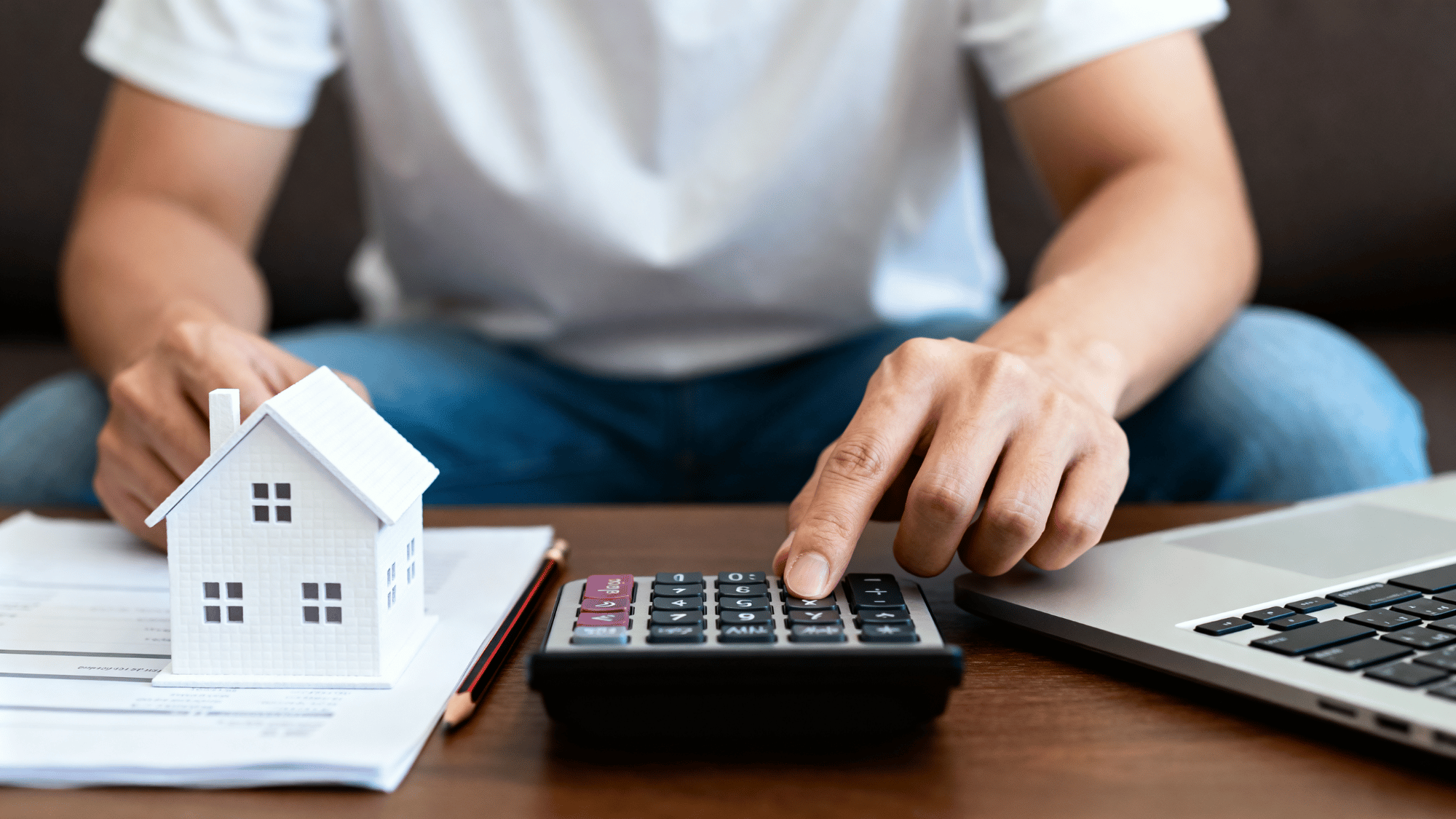 Person calculating mortgage payments with model house, calculator, and laptop on desk home.