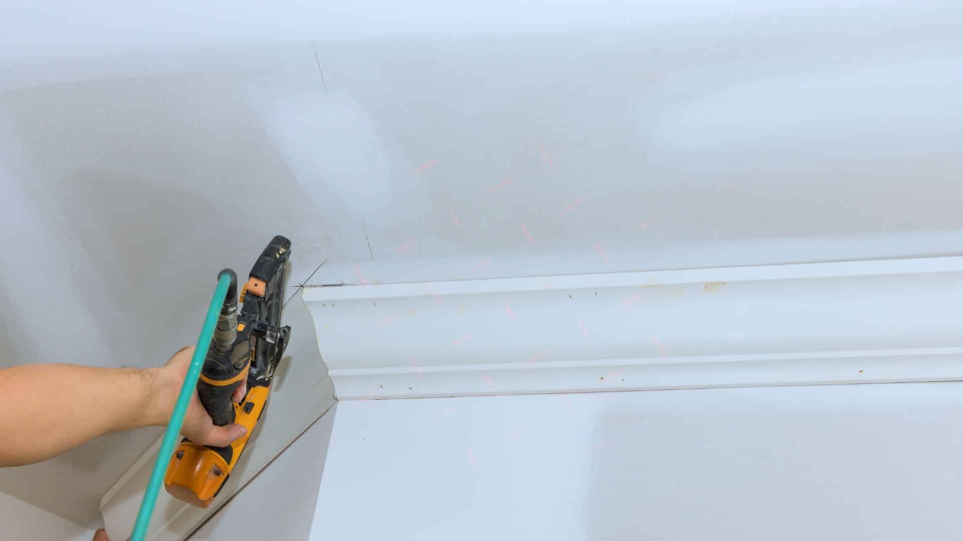 Person using a nail gun to install crown molding, with a laser level aligned to ensure accurate placement on the ceiling