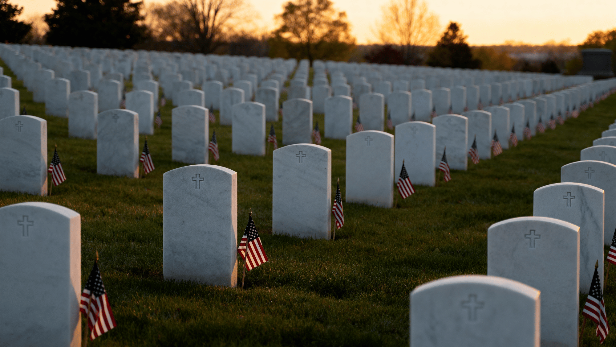 Rows of white headstones with flags at sunrise honoring fallen heroes