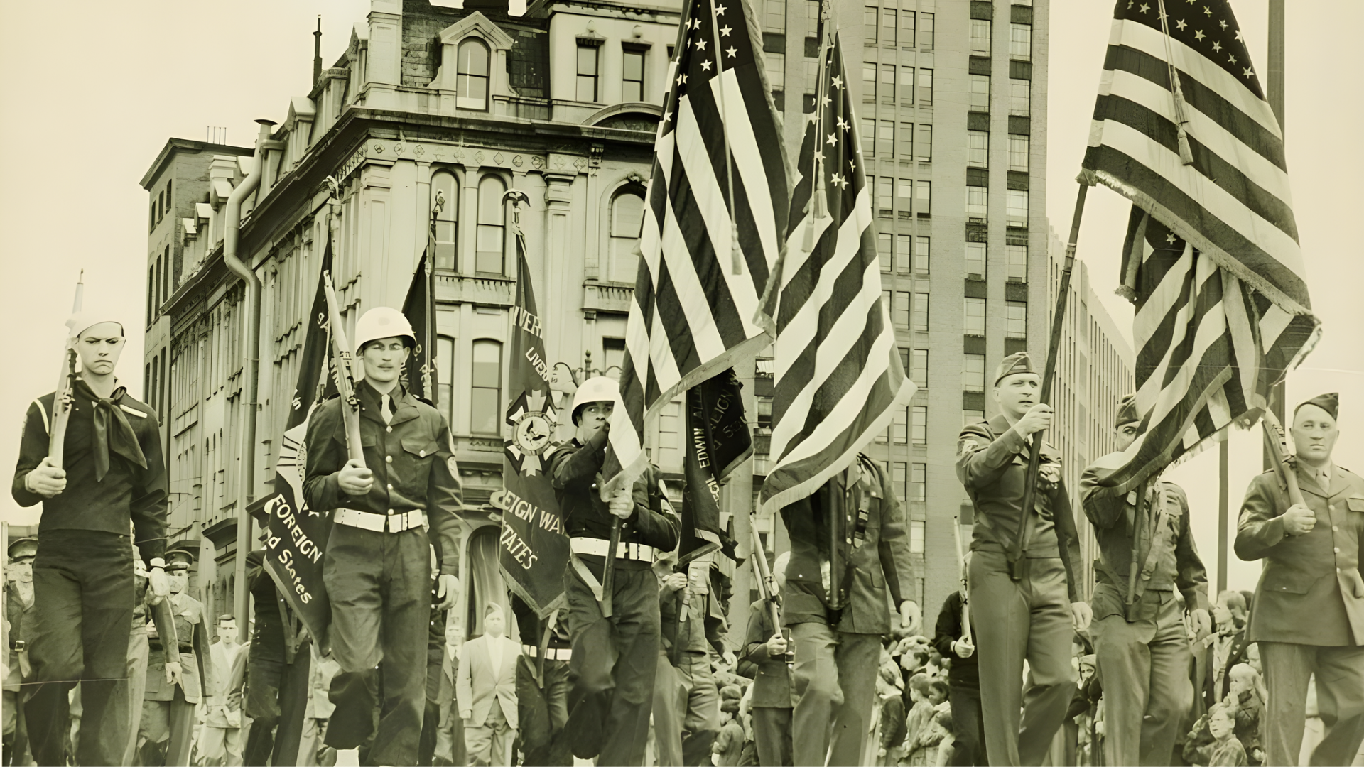 Sepia-toned photo of soldiers and a sailor carrying American flags and banners in a city parade