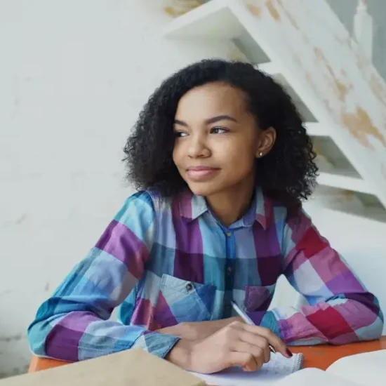 Young woman in plaid shirt writing in notebook at orange table indoors