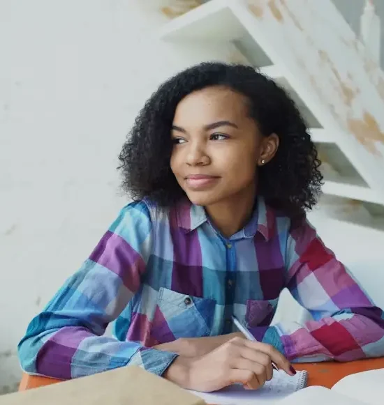 Young woman in plaid shirt writing in notebook at orange table indoors