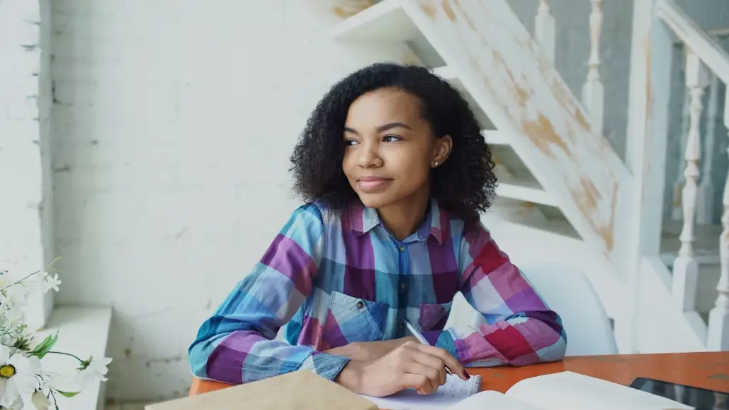 Young woman in plaid shirt writing in notebook at orange table indoors