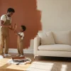 Father and son painting wall terracotta in bright living room with beige sofa and potted plant