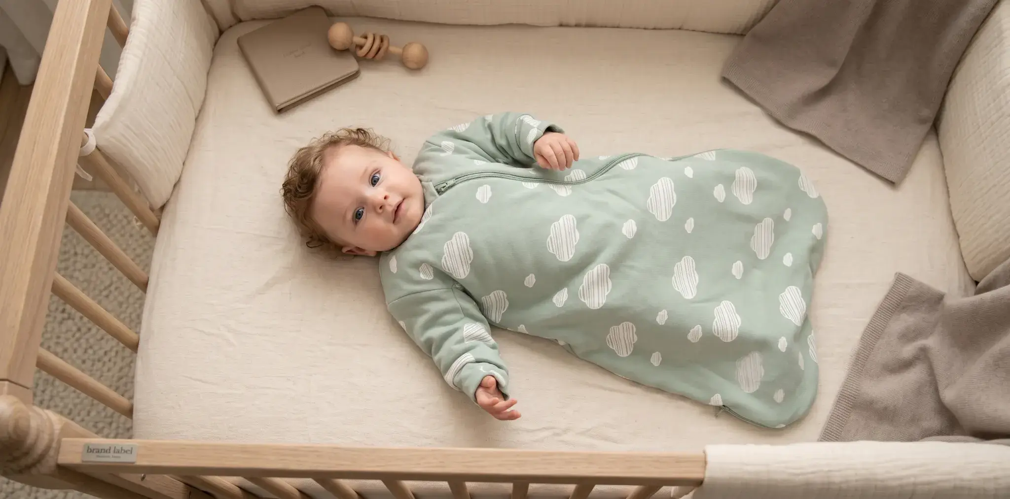 Baby in pale green cloud-patterned sleepsuit lying in a wooden crib with beige bedding