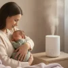 Mother cradling sleeping baby beside humidifier on wooden nightstand in softly lit bedroom