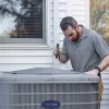 Technician inspecting outdoor air conditioning unit beside house with flashlight