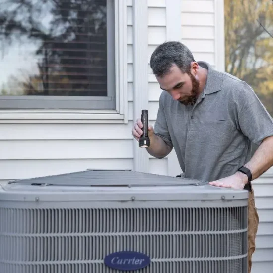Technician inspecting outdoor air conditioning unit beside house with flashlight