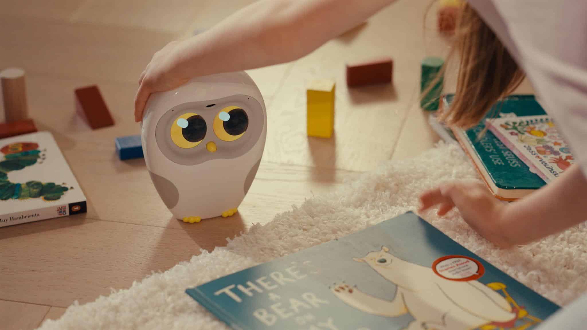 Robotic owl toy with bright eyes on wooden floor surrounded by children's books and blocks