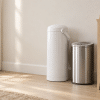 White diaper pail and stainless steel trash can beside wooden dresser in sunlit nursery